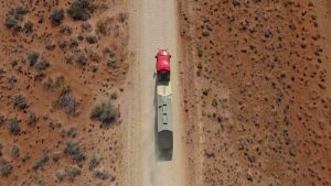 Aerial view of a red Kodiak-equipped truck hauling a trailer along a dusty lease road in a remote desert landscape.