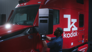 Technician removing a Kodiak SensorPod module from the mirror of a red autonomous truck in a maintenance bay.