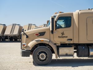 Side view of a tan frac sand truck parked on a yard with multiple sand trailers staged in the background.