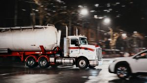 Silver tanker truck traveling along a wet city street at night with reflections on the pavement.