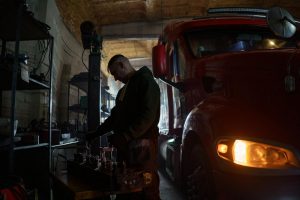 Mechanic working beside a red semi truck in a dimly lit repair shop.