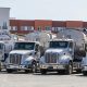 Four polished Peterbilt semi‑trucks with bulk grain trailers parked in an industrial yard.