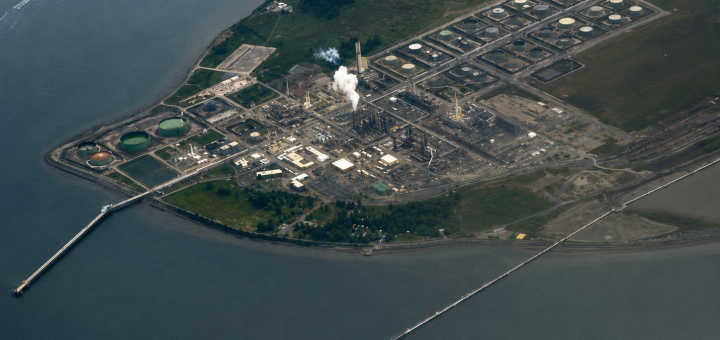 Aerial view of a waterfront refinery and storage tank complex illustrating how the global refinery crunch can affect refined product supply, fuel logistics, and downstream transportation markets.