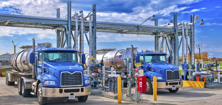 Two tanker trucks positioned at a fuel-loading terminal with overhead piping and transfer equipment.