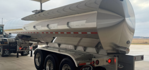 Rear three-quarter view of a polished stainless-steel tank trailer with three axles beneath a building canopy.