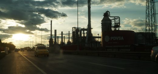 El Palito refinery in Venezuela at sunset, with roadside traffic, industrial towers and a red PDVSA storage tank.