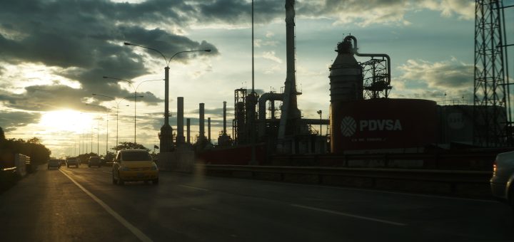 El Palito refinery in Venezuela at sunset, with roadside traffic, industrial towers and a red PDVSA storage tank.