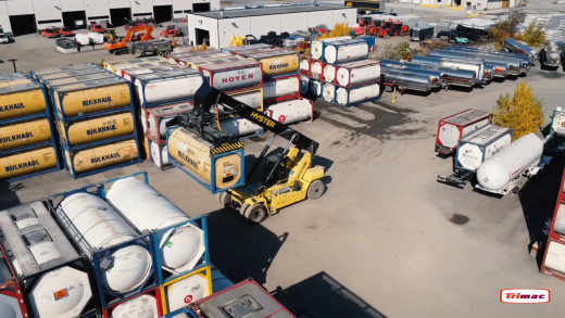 Yellow reach stacker lifting an ISO tank container beside stacked tank containers and silver tank trailers.