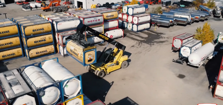Yellow reach stacker lifting an ISO tank container beside stacked tank containers and silver tank trailers.