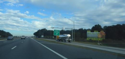 A silver tank trailer enters a weigh-station lane beside a green highway sign on Interstate 75.